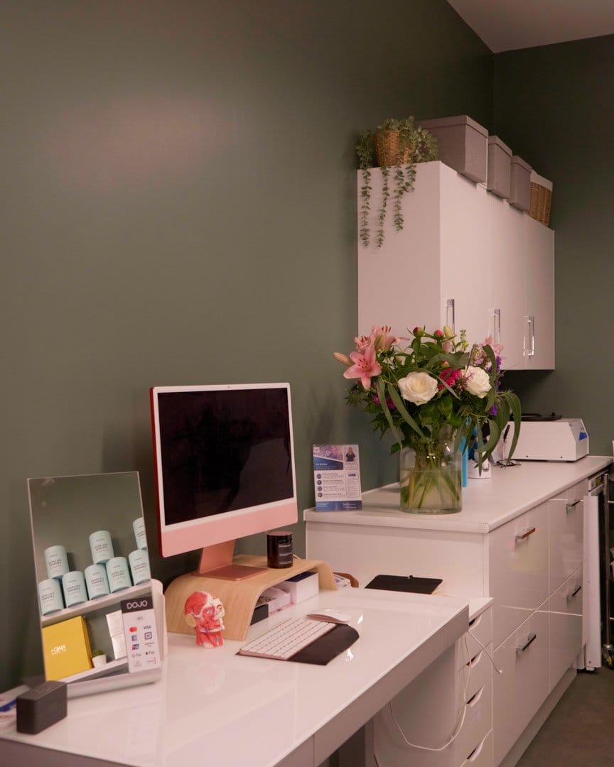 A clinic desk with a pink computer, fresh flowers and a shelf of skincare products